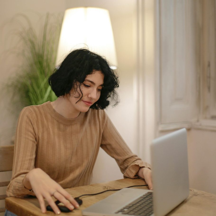 Woman working on a laptop at a wooden table in a well-lit room.