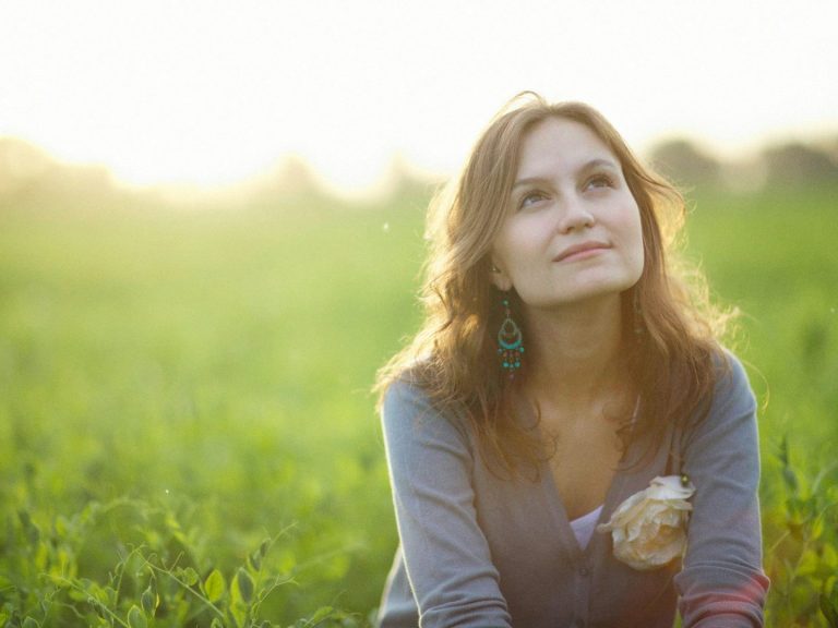 Smiling woman outdoors, looking up thoughtfully in a green field with soft sunlight.