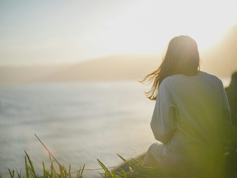 Person sitting by the sea, gazing at the sunset with flowing hair.
