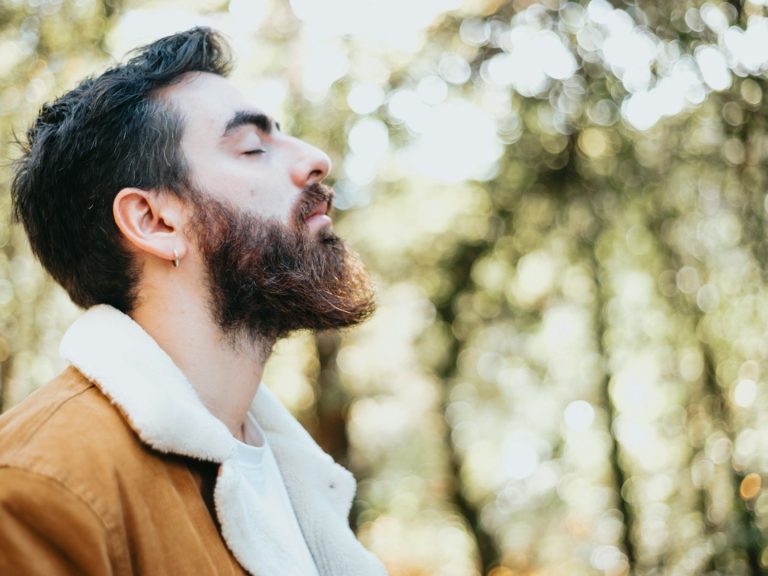 A man with a beard stands outdoors, breathing deeply amidst trees.