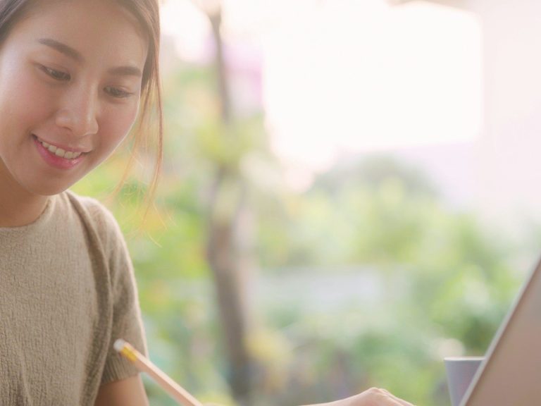 Woman smiling while writing in a notebook beside a laptop in a bright, natural setting.